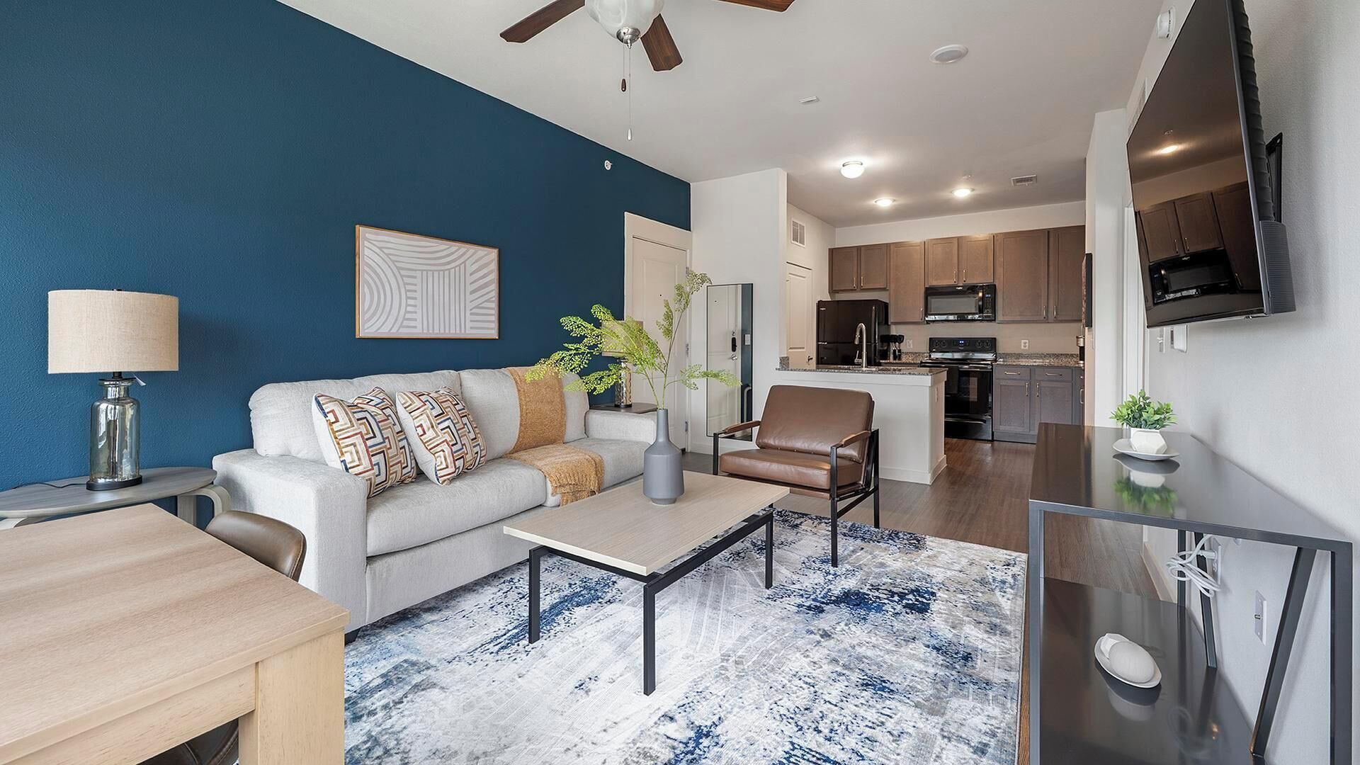 A modern living room with a sofa, coffee table, and a TV. There is a kitchen area with cabinets and appliances in the background.