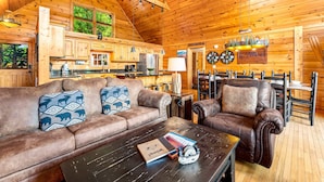 Accogliente interno di una baita in legno con soffitto in legno, un divano in pelle con cuscini decorati, un tavolino da caffè con un libro e un oggetto decorativo, e un'area pranzo con tavolo e sedie in legno.