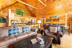 A cozy log cabin interior with a wooden ceiling, a leather sofa with patterned cushions, a coffee table with a book and a decorative item, and a dining area with a wooden table and chairs. - Luxury Mtn Retreat W Breathtaking Views, Sleeps 14 (Gatlinburg)