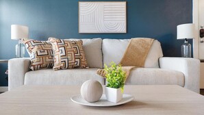 Een woonkamer met een bank, gestreepte kussens, een vaas met groene planten en een rond, textuurrijk object op een tafel.