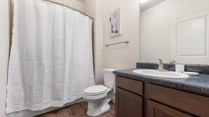 A bathroom with a white toilet, a shower curtain, a sink with a granite countertop, and a towel rack. - Landing | Chic 2BD, Gym, Pool (Lockhart)