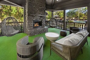Une terrasse extérieure dotée d’une cheminée en pierre, de meubles en osier et offrant une vue sur une piscine et des arbres.