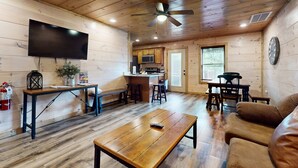 A cozy cabin interior with a wooden floor, a ceiling fan, a flat-screen TV, a kitchen area, and a seating area with a couch and chairs. - 4BR Cabin Near Dollywood・2 Hot Tubs・Grill・Pool (Pigeon Forge)