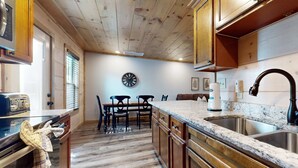 A kitchen with wooden cabinets, a granite countertop, a stainless steel sink, and a dining area with a table and chairs. - 4BR Cabin Near Dollywood・2 Hot Tubs・Grill・Pool (Pigeon Forge)