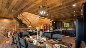 A spacious wooden cabin with a dining area set for four, a kitchen with modern appliances, and a staircase leading to an upper level. - Spacious Scenic Cabin, Sleep 12, Games, Hot Tub, View (Sevierville)