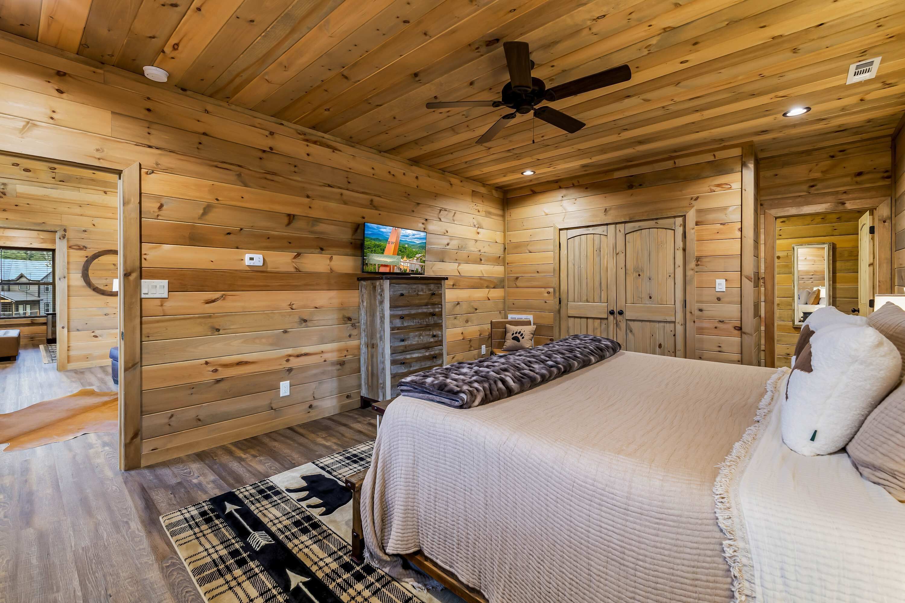 A wooden cabin bedroom with a bed, a ceiling fan, a fireplace, and a television.