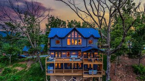 A multi-story wooden house with a blue roof, surrounded by trees and a forested area. - Spacious Scenic Cabin, Sleep 12, Games, Hot Tub, View (Sevierville)