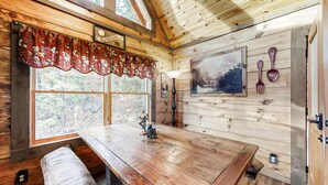 Interno di una baita in legno con un tavolo, una panca e una finestra decorata con un tendaggio floreale.
