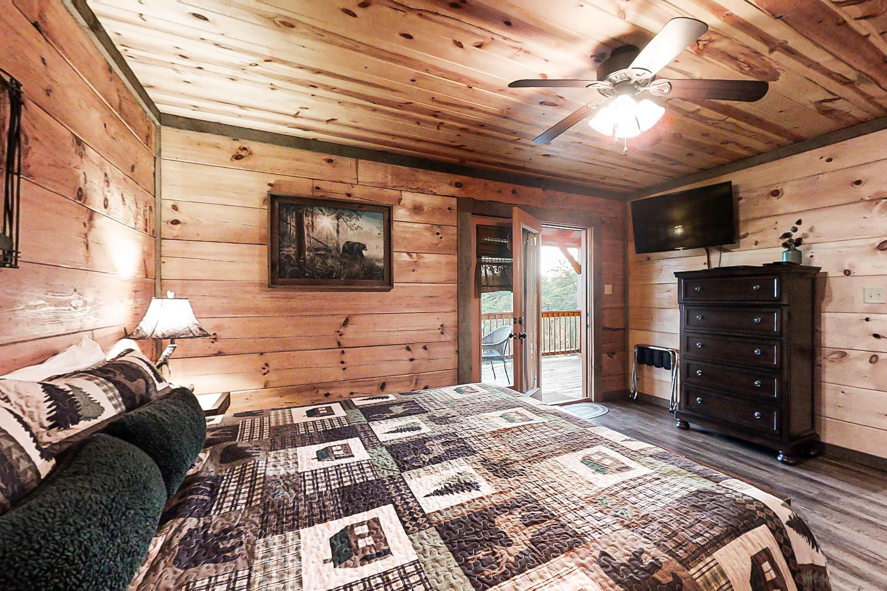 Quarto de cabana de madeira com cama acolchoada, ventilador de teto, televisão e cómoda.