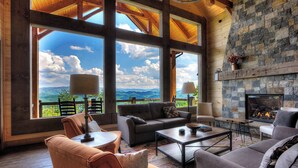 A spacious living room with large windows, a stone fireplace, and comfortable seating. - Cozy Cabin W Home Theater, Sleeps 15, Hot Tub, Views (Sevierville)