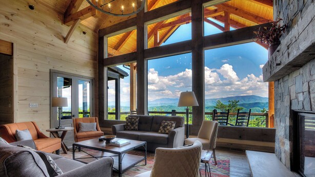 A spacious living room with large windows offering a mountain view, wooden beams on the ceiling, and a stone fireplace. - Cozy Cabin W Home Theater, Sleeps 15, Hot Tub, Views (Sevierville)