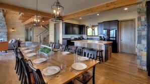 A spacious dining area with a long wooden table set for a meal, flanked by dark wooden chairs. A modern kitchen with dark cabinets and stainless steel appliances is visible in the background. - Cozy Cabin W Home Theater, Sleeps 15, Hot Tub, Views (Sevierville)