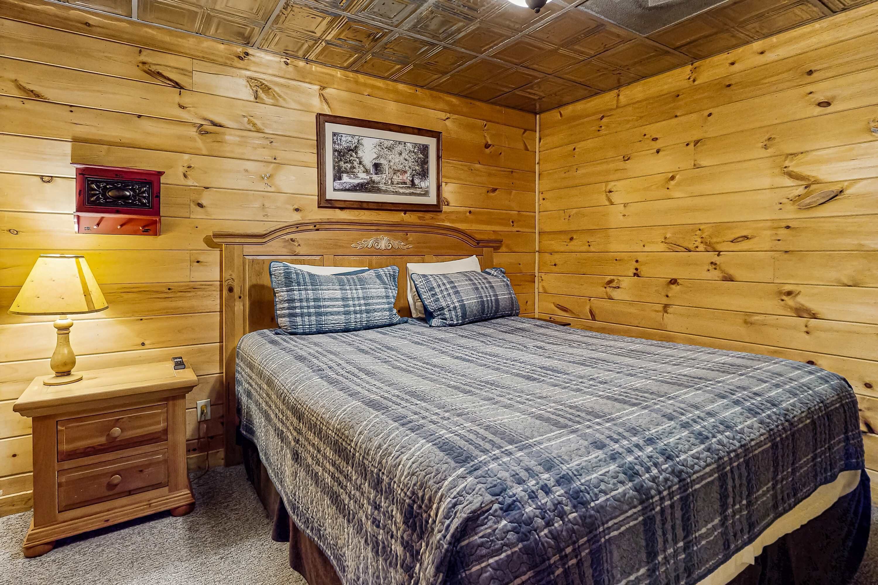 A wooden-paneled room with a bed, a nightstand with a lamp, and a framed picture on the wall.