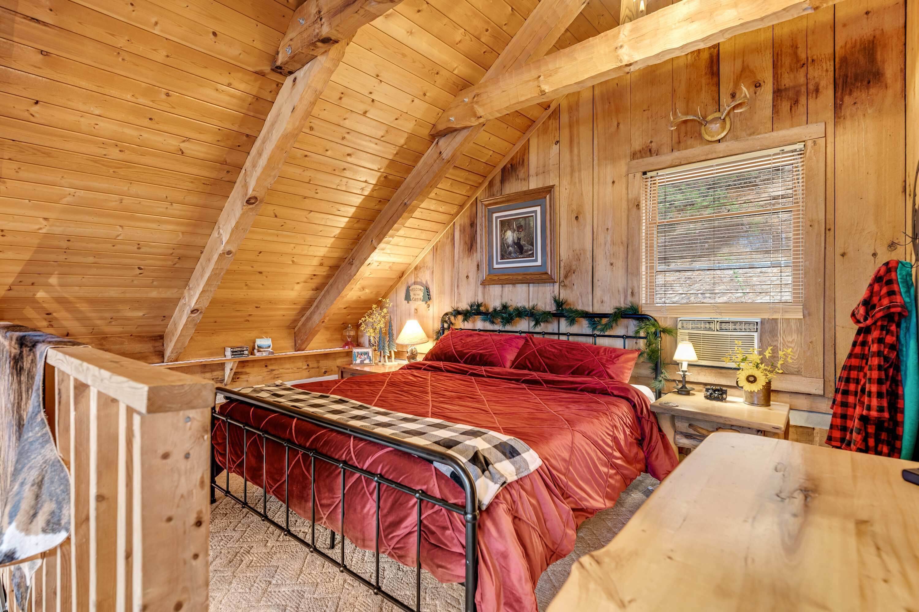 Une chambre confortable située sous les toits, avec un plafond en bois, un lit recouvert d’une literie rouge, une table de chevet avec une lampe et une fenêtre équipée de stores.