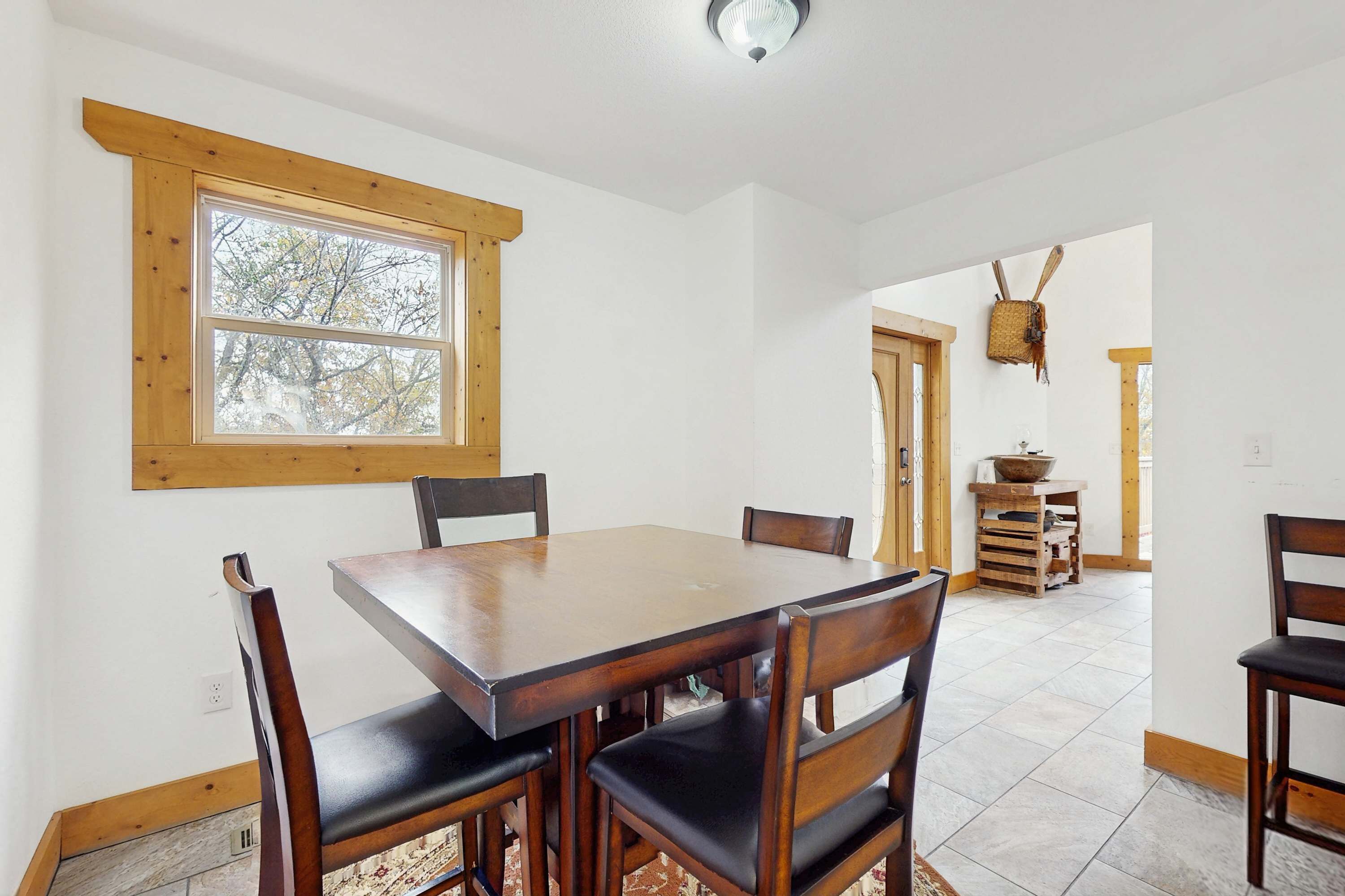 A dining area with a wooden table and chairs, a window with a view of trees, and a kitchen area in the background.