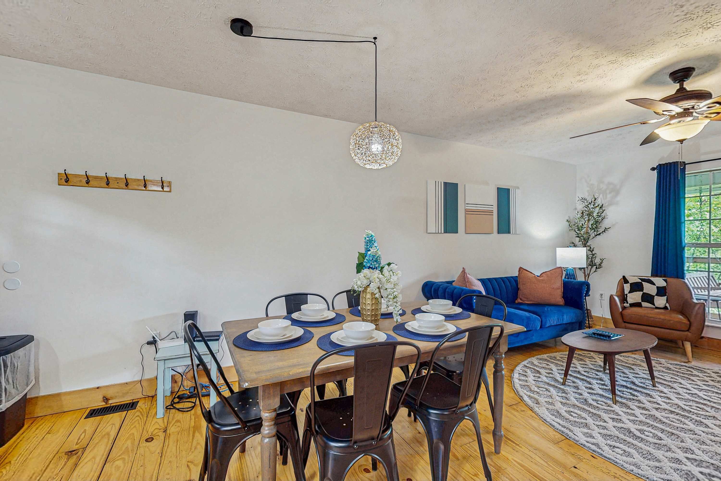 A dining area with a wooden table set for four, surrounded by chairs. There is a ceiling fan, a hanging light fixture, and a coat rack on the wall.