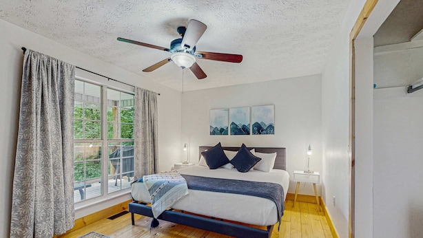 Une chambre à coucher comprenant un lit, un ventilateur de plafond, une table de chevet, une fenêtre avec des rideaux et une vue sur la verdure à l’extérieur.