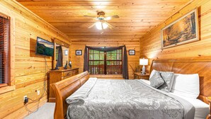 A wooden cabin bedroom with a bed, nightstand, TV, and a large window with curtains. - Always Delightful - Sleeps 10, Hot Tub, Fire pit, Games (Sevierville)