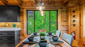 A wooden cabin interior with a dining table set for six, a kitchen area, and a large window offering a view of greenery. - The Treehouse, Sleep 12・Hot Tub・Firepit・Game Room (Sevierville)