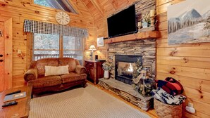 A cozy log cabin interior with a stone fireplace, a couch, a television, and a window with blinds. - 3.5 Mi to DT Pigeon Forge・Dog Ok! Getaway・Hot Tub (Pigeon Forge)
