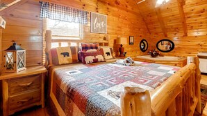 A cozy log cabin bedroom with a wooden ceiling, a bed with a quilt, a nightstand with a lantern, and a window with checkered curtains. - 3.5 Mi to DT Pigeon Forge・Dog Ok! Getaway・Hot Tub (Pigeon Forge)