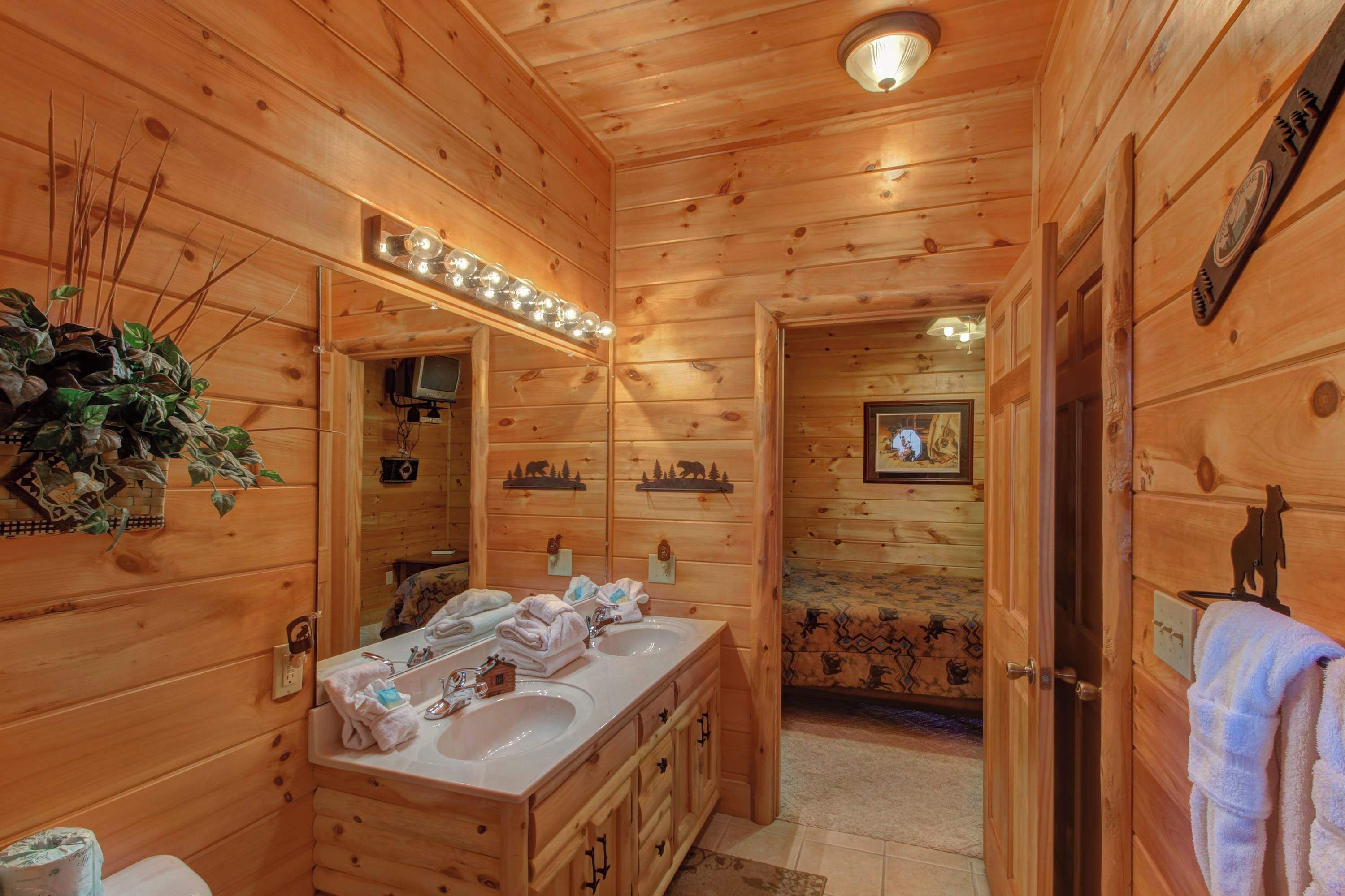 A wooden cabin bathroom with a double sink vanity, a mirror with lights, a toilet, and a door with a bear decoration.