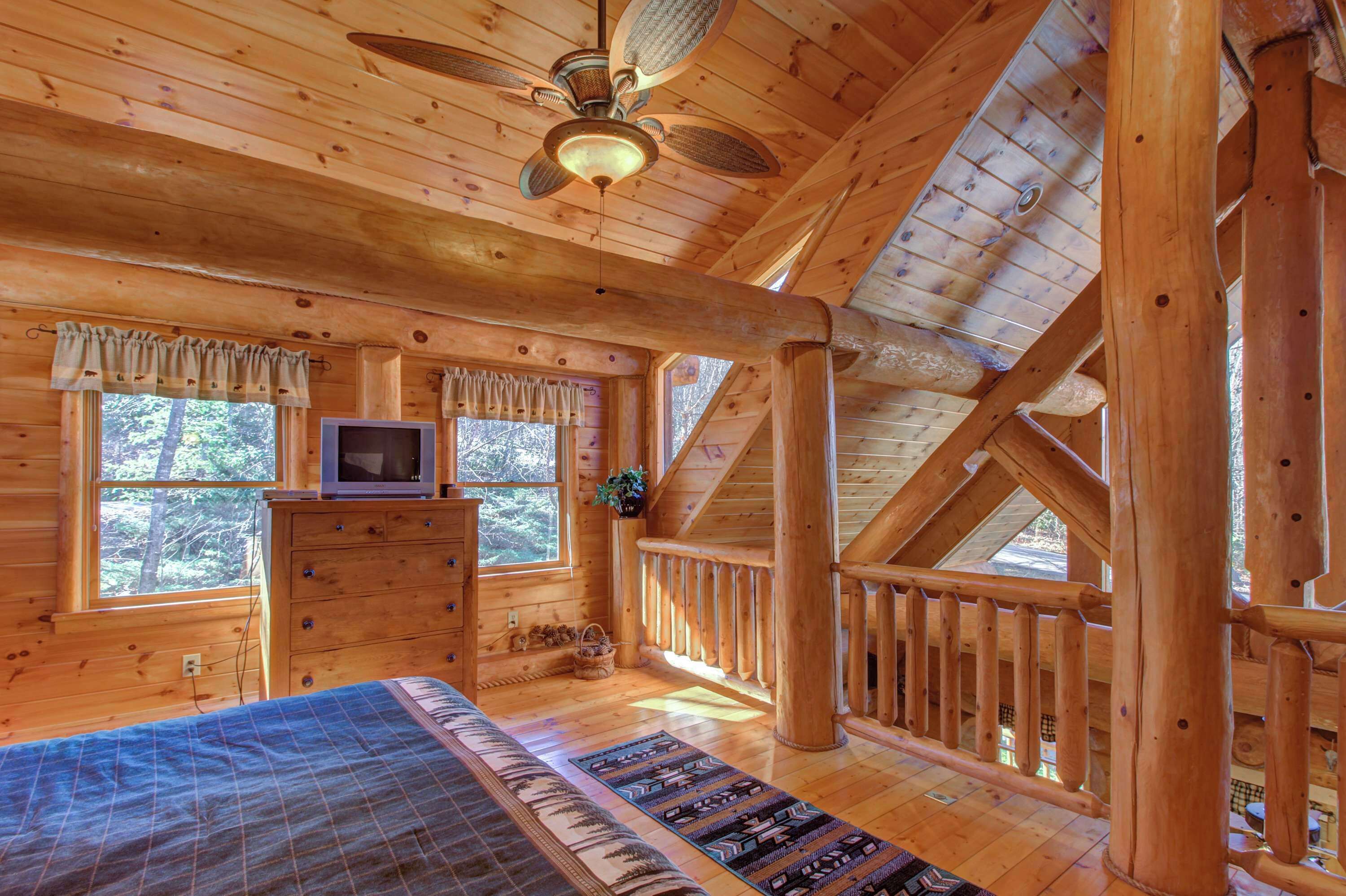 A log cabin room with a bed, dresser, TV, and windows overlooking a forest.