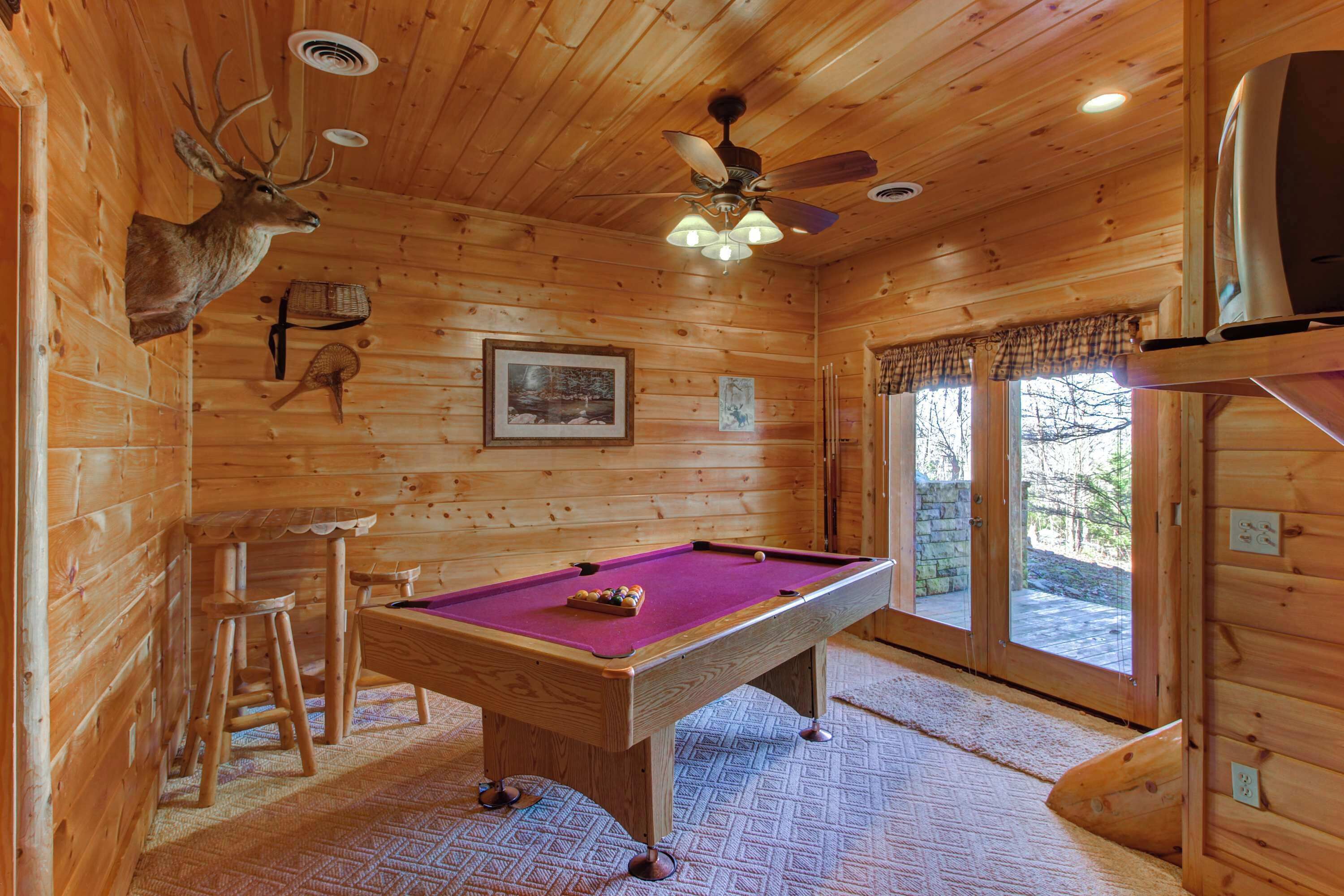 A wooden cabin interior with a pool table, a ceiling fan, and a mounted deer head.