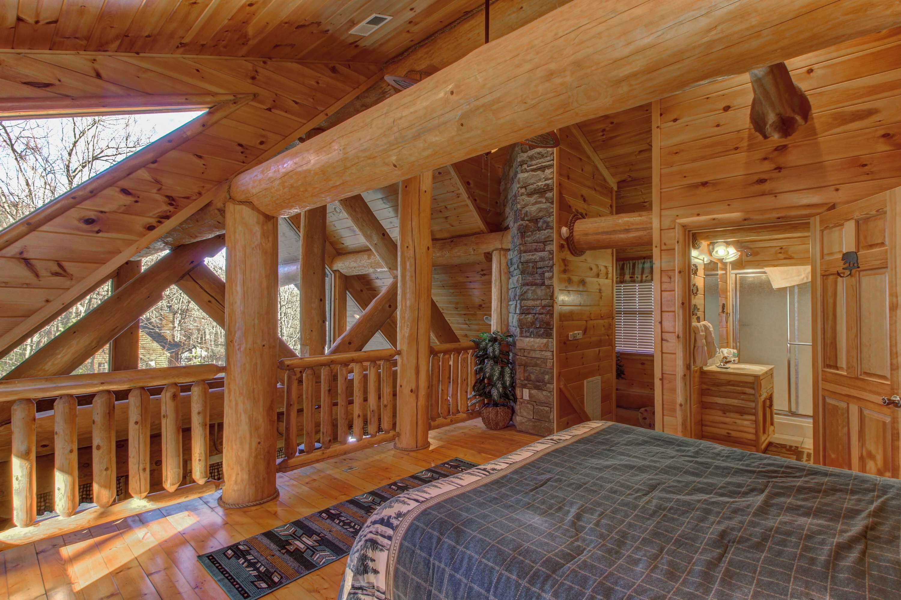 A log cabin bedroom with a large bed, wooden beams, a stone fireplace, and a balcony with a railing.