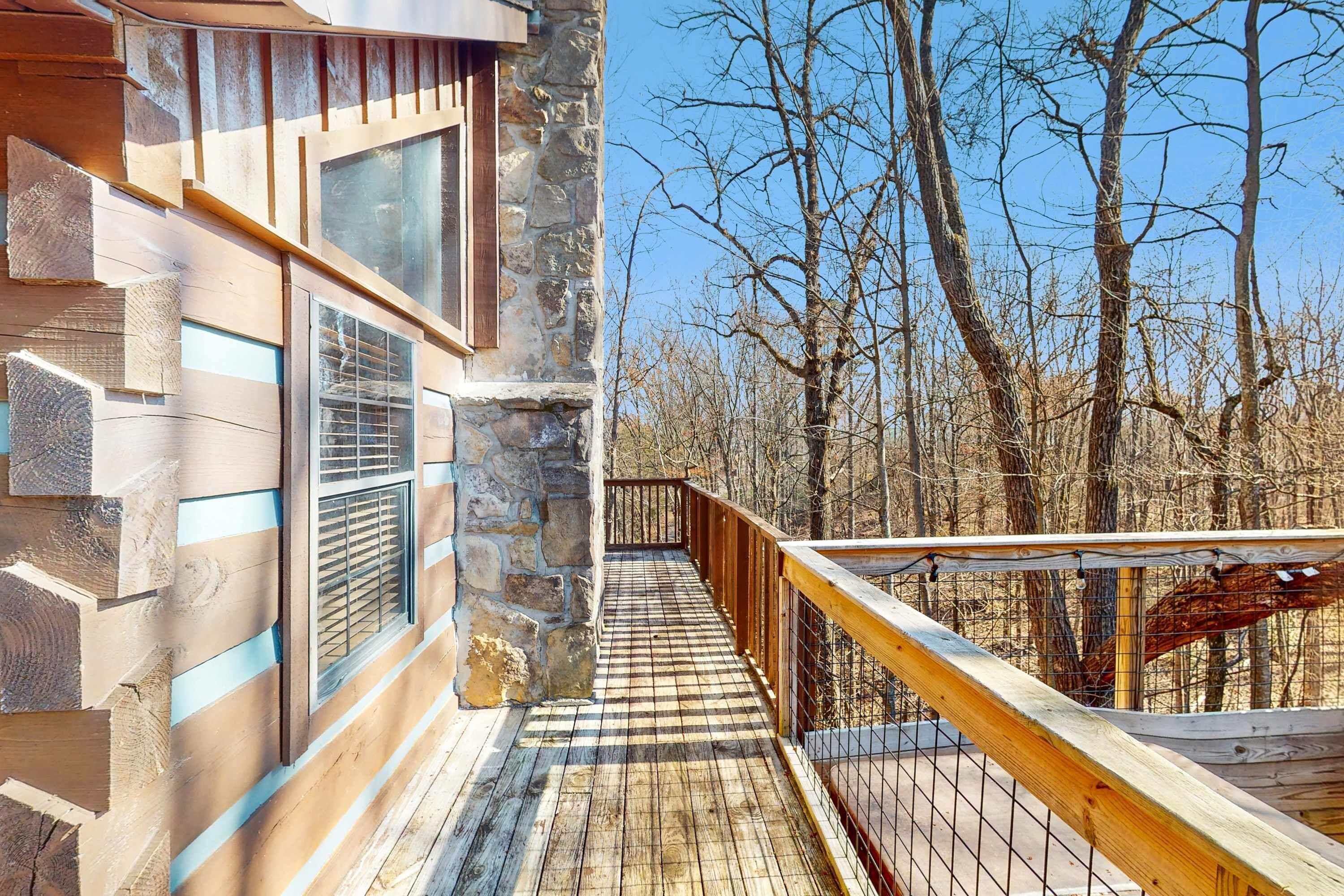 Une terrasse en bois, avec un mur en pierre et une rambarde en métal, qui mène à une zone boisée.