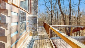 Une terrasse en bois, avec un mur en pierre et une rambarde en métal, qui mène à une zone boisée.