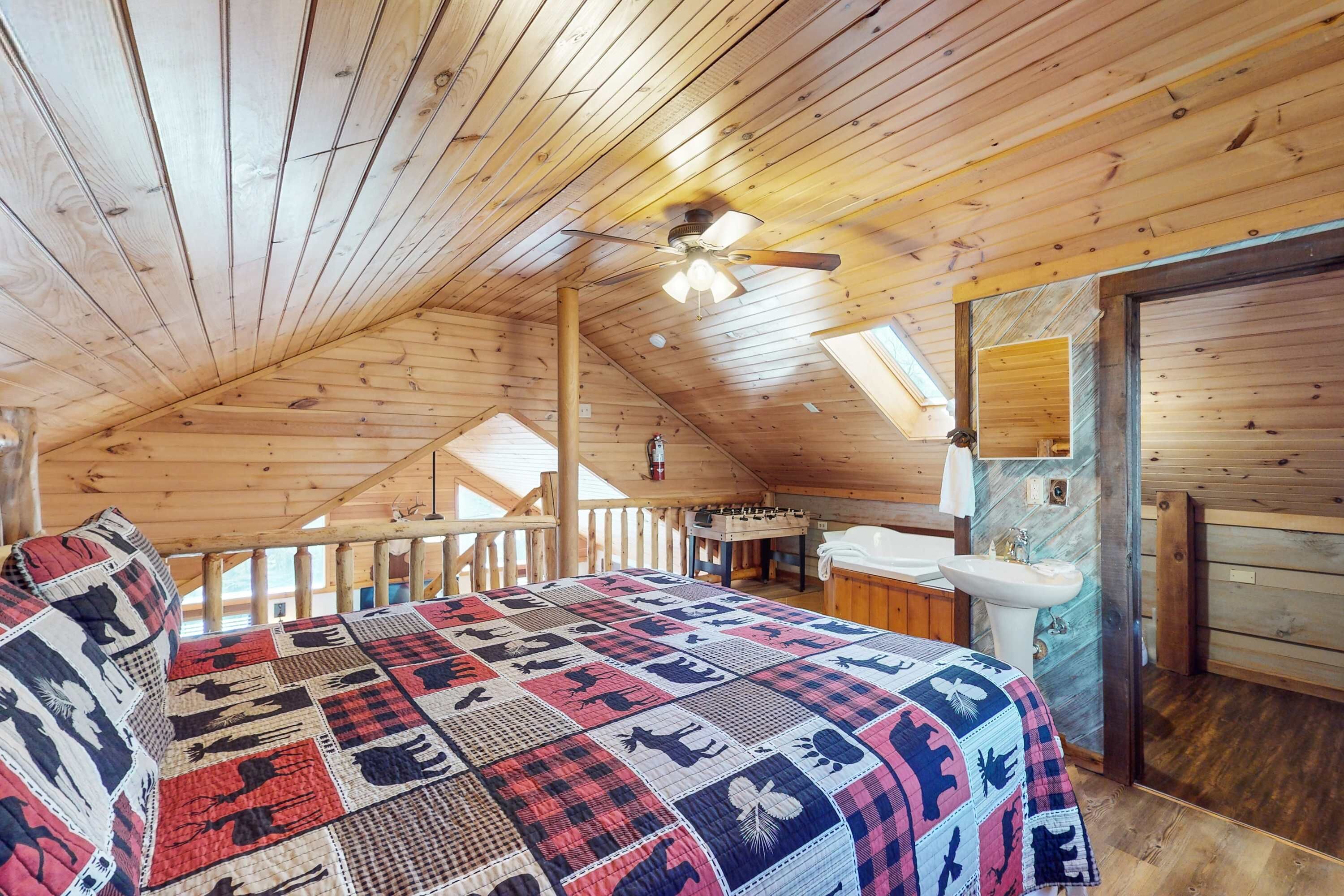 Une chambre confortable située dans les combles, avec un plafond en bois, un lit recouvert d’une courtepointe, un ventilateur de plafond et une salle de bain équipée d’un lavabo et d’un miroir.