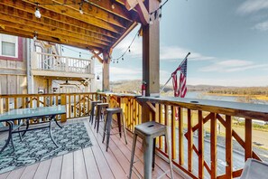 Una terraza de madera con una mesa de vidrio y taburetes, con vistas a un paisaje pintoresco.