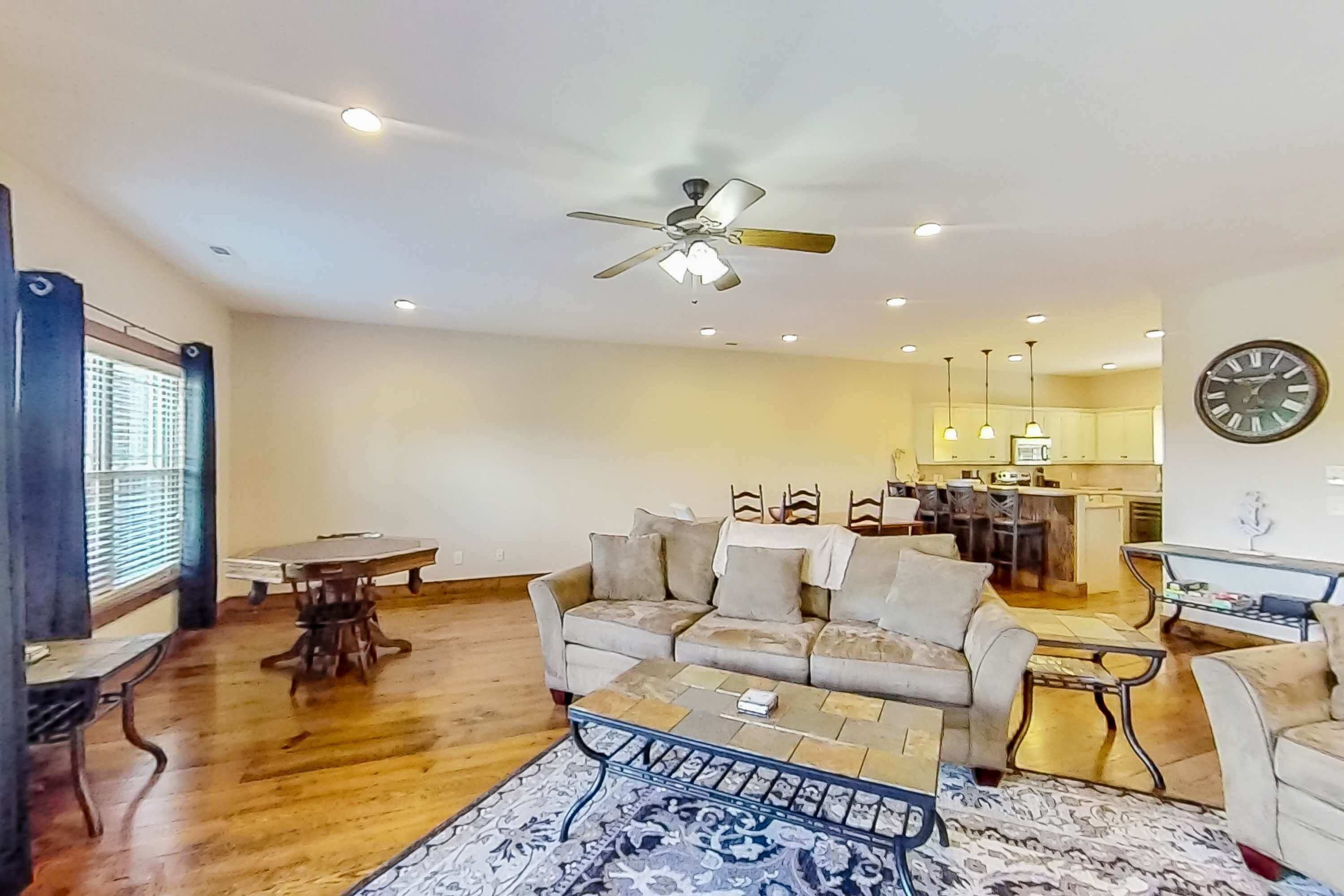 A spacious living room with a ceiling fan, a dining area with a table and chairs, and a kitchen in the background.