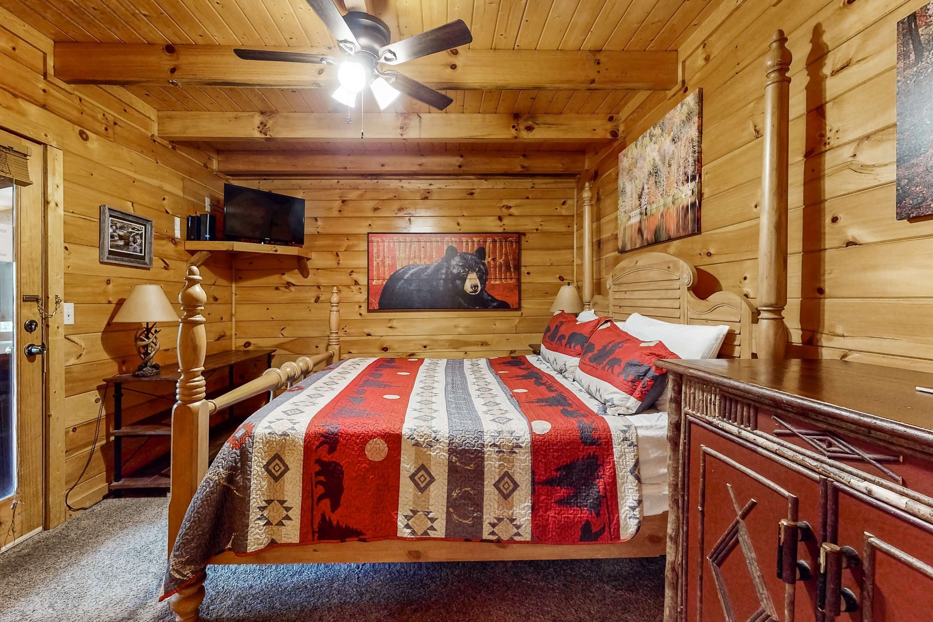 Een slaapkamer in een houten chalet met een bed, een plafondventilator, een tv aan de muur en een schilderij van een beer.