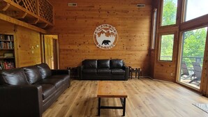 A wooden interior with a logo on the wall, a leather sofa, a wooden coffee table, and a bookshelf. - 13.7 Mi to DT  Gatlinburg・Spacious Cabin・Hot Tub (Gatlinburg)