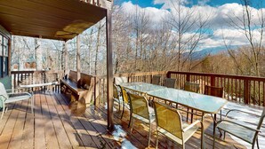 Eine Holzterrasse mit einem Glastisch, Stühlen, einer Bank und Blick auf Bäume und Berge.