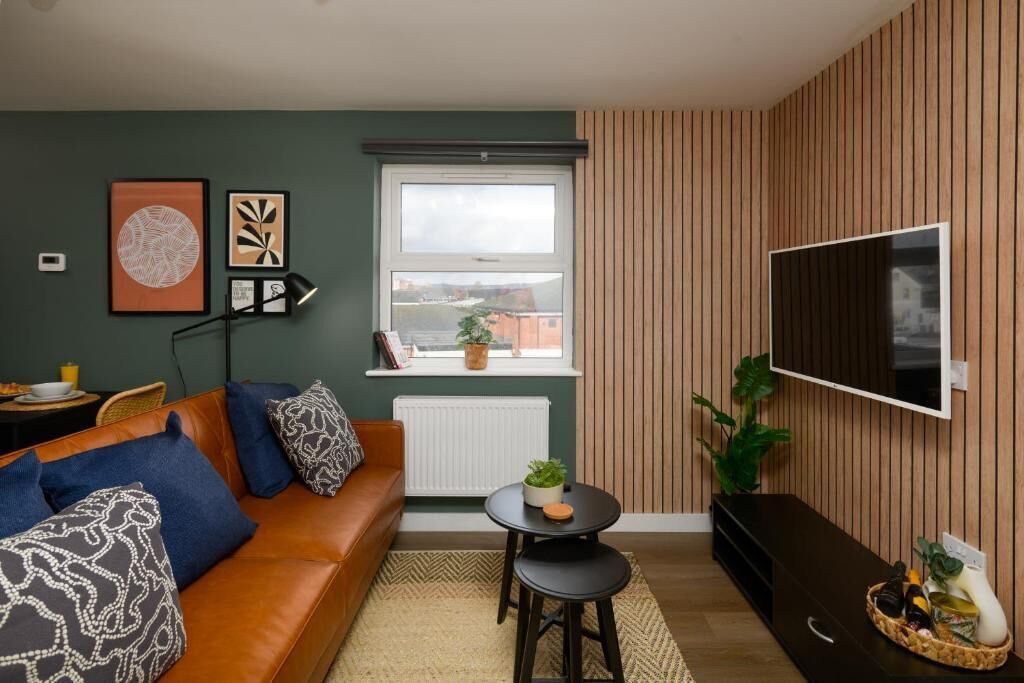 A modern living room with a brown leather sofa, a flat-screen TV, a small round table with a plant, and a window with a view of buildings.