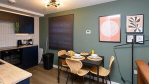 A compact kitchen with a dining area, featuring a modern design with a dark wall, a ceiling light, and framed artwork. - Benjamin Suite Modern 3BR Apartment (Cardiff)