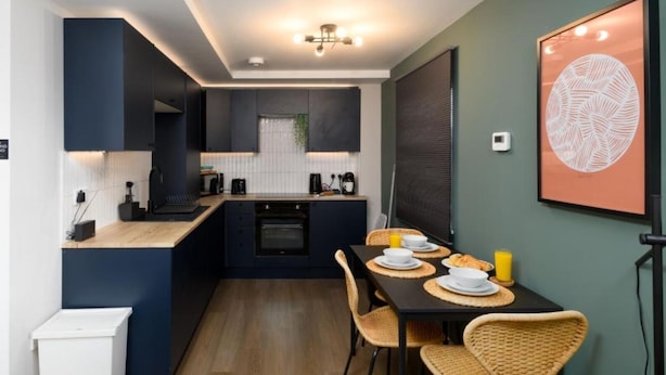 A compact kitchen with dark cabinetry, a central island, and a dining area with wicker chairs. - Benjamin Suite Modern 3BR Apartment (Cardiff)