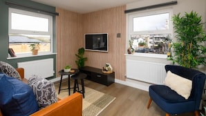 A modern living room with a TV mounted on the wall, a comfortable sofa, a blue armchair, and a small dining area. - Benjamin Suite Modern 3BR Apartment (Cardiff)