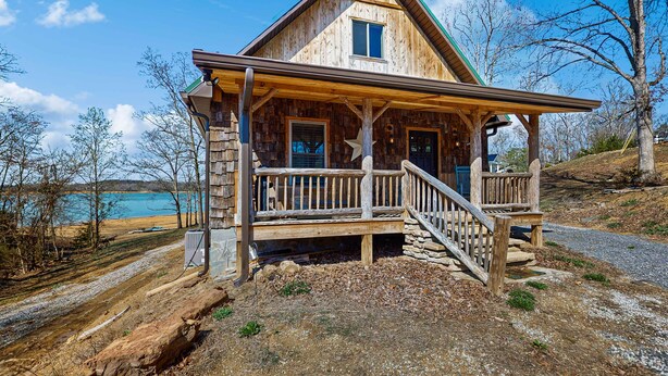 A wooden cabin with a porch and a stone foundation, located near a body of water. - Cozy Lake Front Cabin, Sleeps 4, Dock, Play Yard (Dandridge)