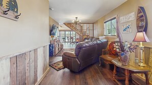 A spacious living room with a leather sofa, a wooden coffee table, and a chandelier. A staircase leads to an upper level, and there are framed pictures on the walls. - Cozy Lake Front Cabin, Sleeps 4, Dock, Play Yard (Dandridge)