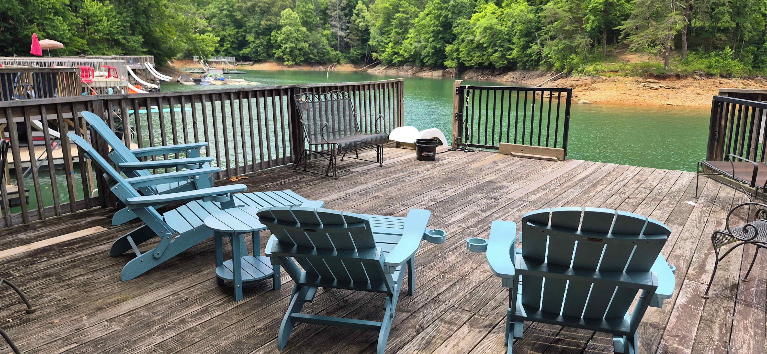 A wooden deck with Adirondack chairs and a table, overlooking a body of water with a dock and boats in the background.