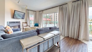 A modern living room with a grey sectional sofa, a flat-screen TV mounted on the wall, a fireplace, and large windows with curtains. - Riverfront Retreat・1 mile to Pigeon Forge・Pool (Pigeon Forge)