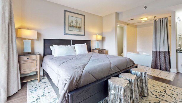 A hotel room with a large bed, two bedside tables with lamps, a painting on the wall, a bathroom with a bathtub, and a shower curtain. - Riverfront Retreat・1 mile to Pigeon Forge・Pool (Pigeon Forge)