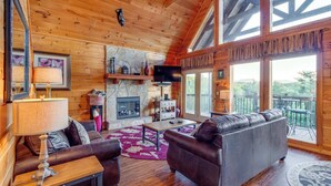 A cozy cabin interior with a fireplace, leather sofas, wooden floors, and large windows offering a view of the outdoors. - Spacious, Dog Welcome Cabin, Sleeps 8・Mtn Views (Pigeon Forge)