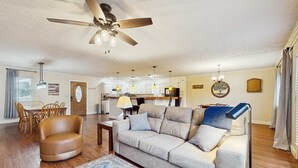 Ein Wohnzimmer mit einer Couch, einem Couchtisch, einem Esstisch mit Stühlen, einem Deckenventilator, Pendelleuchten und einer Küche im Hintergrund.