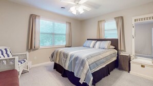 Um quarto com uma cama, uma cadeira, uma mesa de cabeceira, um ventilador de teto e uma janela com cortinas.