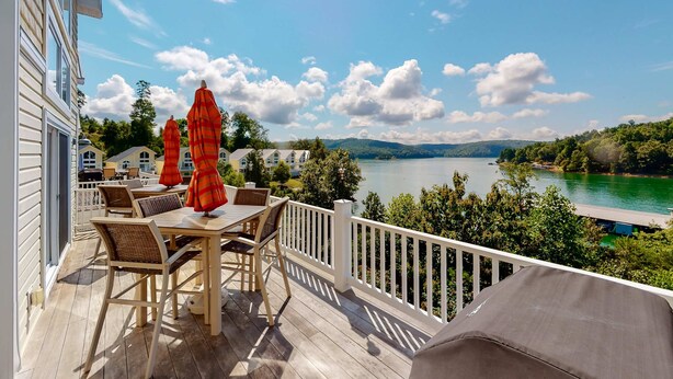 Een houten terras met buitenmeubilair en parasols, met uitzicht op een meer en heuvels.